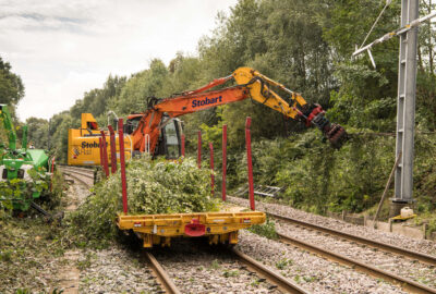Stobart Rail excavator on a railway flatbed clearing overgrown branches and foliage along the tracks.