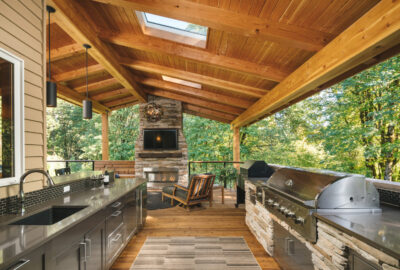 Outdoor covered kitchen and living area with stone fireplace, stainless steel grill, and wooden vaulted ceiling.