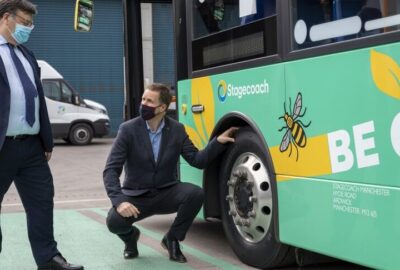 Two men in suits and masks inspect a green Stagecoach bus; one man crouches by the wheel.