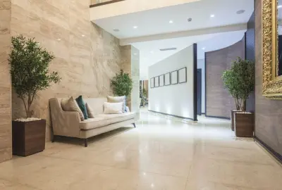 Modern hotel lobby with a beige sofa, potted plants, marble walls, and a long brightly lit hallway.