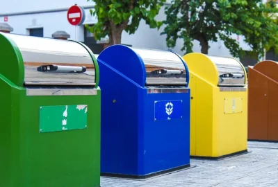 Row of four colorful recycling bins in green, blue, yellow, and brown on a sidewalk.