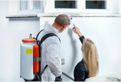 A worker in a white hazmat suit and sprayer backpack looks at a wall while a woman points to a spot.