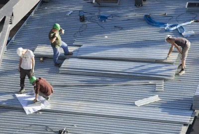 Four construction workers installing corrugated metal roofing sheets on a large rooftop.