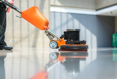 Person operating an orange industrial floor polisher on a highly reflective floor.