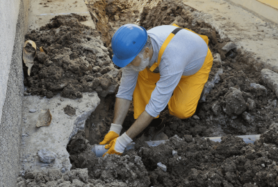 *   Image content: A construction worker in a blue hard hat, white shirt, and yellow overalls is working in a dirt trench. He