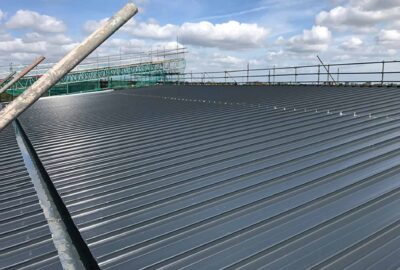 Grey corrugated metal roof under construction with scaffolding in the background under a cloudy sky.