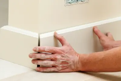 Hands installing white baseboard molding along a corner of a beige wall.