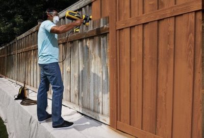 *   Image content: A man wearing safety gear (mask, goggles) is using a yellow paint sprayer to apply brown stain to a wooden