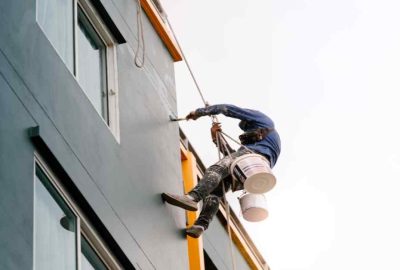 Worker rappelling down the side of a building, painting the exterior wall grey.