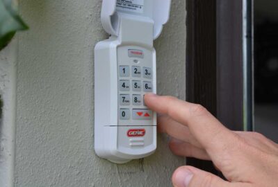 A finger pressing a button on a white Genie numeric keypad mounted on an exterior wall.