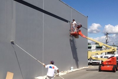 Workers painting a large gray exterior wall, one with a long roller and another in an aerial lift.