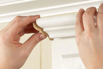 Hands peeling a decorative brown trim piece onto the corner of a white wooden molding.
