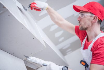 A construction worker in a red cap and safety goggles applying joint compound to a drywall ceiling with a trowel.