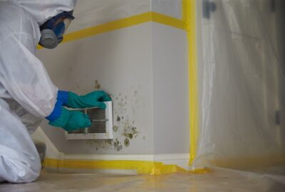 Professional in a hazmat suit and respirator removing a wall vent surrounded by mold growth.