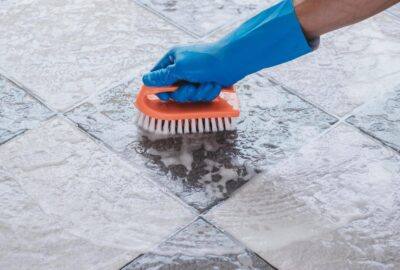 A hand in a blue glove scrubbing a soapy tile floor with an orange scrub brush.