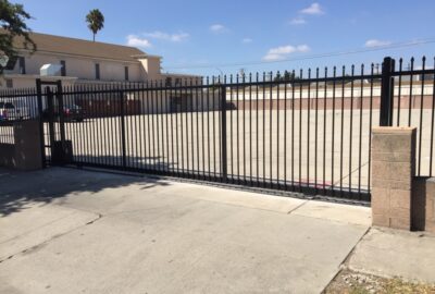 Black metal security gate blocking an empty paved lot in front of a tan building under a clear sky.