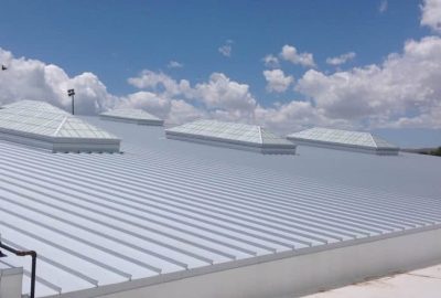 Wide view of a white corrugated metal roof with several skylights under a blue sky with clouds.