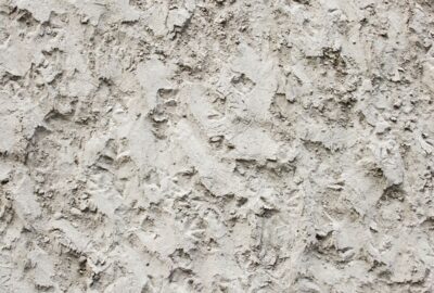 Rough, light gray textured wall with uneven, stucco-like plaster and craggy surface details.