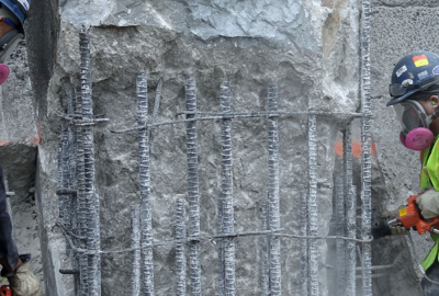 Construction workers in safety gear and respirators chip concrete away from exposed rebar.