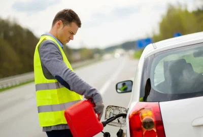 *   Image: A man in a high-visibility yellow vest is pouring fuel from a red gas can into a white car parked on the side of a