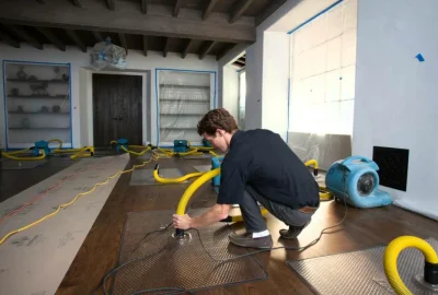 A technician setting up industrial fans and yellow hoses to dry a wooden floor during water damage restoration.