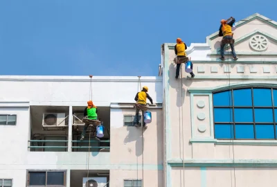 Four painters suspended by safety ropes paint the exterior facade of a multi-story building against a clear blue sky.