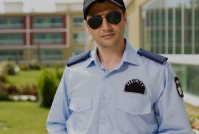Security guard in a light blue uniform and sunglasses holding a walkie-talkie outside a building.