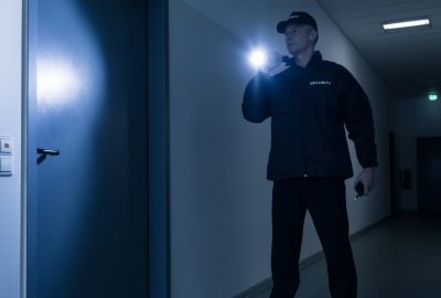 Security guard in a dark, blue-lit hallway holding a flashlight near a blue door.