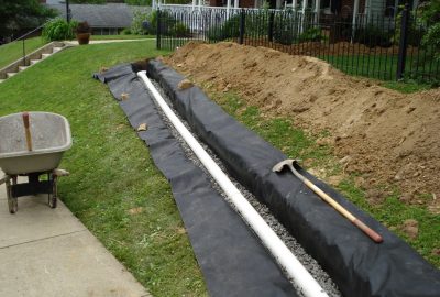 French drain installation with a white perforated pipe in a gravel trench lined with black landscape fabric.