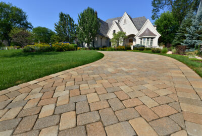 Curved paver driveway leading to a large white luxury home with a manicured lawn and landscaped gardens.