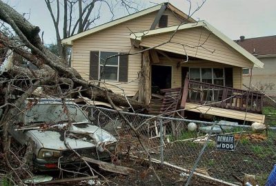 *   Image content: A severely damaged small yellow house, a crushed white car, a fallen large tree, a chain-link fence, and a