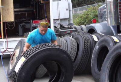 Worker in a blue t-shirt and yellow goggles handling large truck tires next to service vehicles.