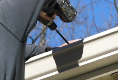 A person using a cordless drill to install or repair white gutters on a house under a blue sky.