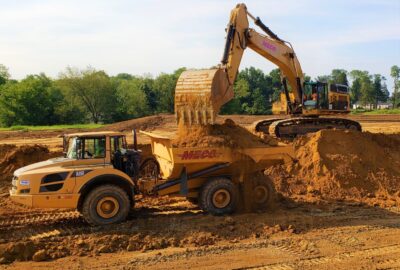 A yellow excavator pours dirt into the bed of a yellow dump truck at a construction site.