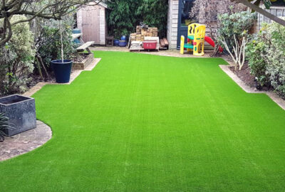 Bright green artificial grass lawn in a backyard with brick borders, garden planters, and a children's play set.