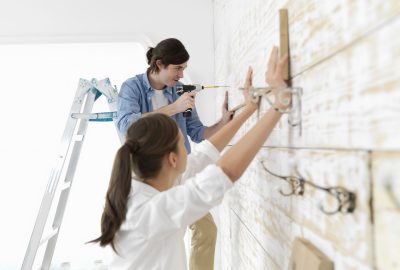 A man uses a power drill to install a wooden shelf on a white paneled wall while a woman holds it in place.