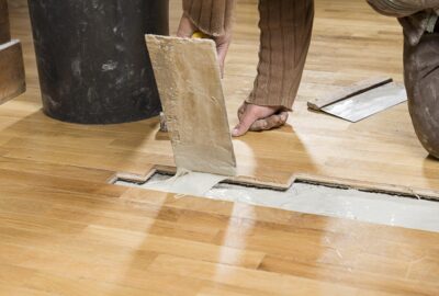 A worker uses a trowel to spread adhesive into a gap between wooden floorboards during installation.