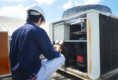 Technician in a hard hat and blue uniform checking an outdoor air conditioning unit with a manifold gauge.