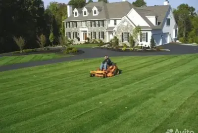A person using an orange ride-on mower on a large striped lawn in front of a large white house.