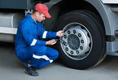 Mechanic in blue overalls and red cap checking a truck's tire pressure with a digital gauge.