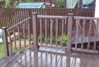 Wet dark brown composite decking and railings in a backyard with a wooden picket fence in the background.