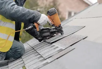 A worker in a high-visibility vest uses a pneumatic nail gun to install grey shingles on a sloped roof.