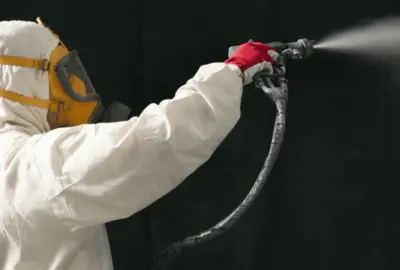 Person in a white hazmat suit and yellow respirator spraying a fine mist with a handheld spray gun.
