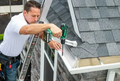 A man on a ladder uses a cordless drill to install white gutters on a house with grey shingles.