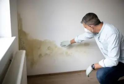 Man in a mask and gloves inspecting water damage and mold on a white wall.