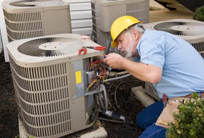 HVAC technician in a yellow hard hat using a screwdriver to repair the wiring of an outdoor air conditioning unit.
