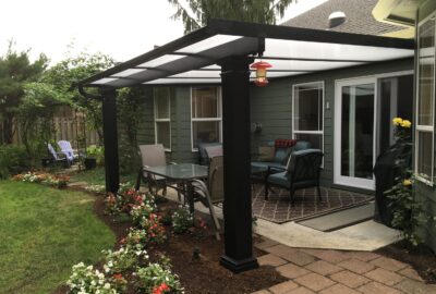 A backyard patio with a black pergola, dining table, lounge seating, and flower beds next to a green house.