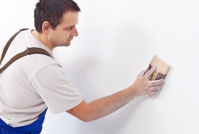 Man in blue overalls sanding a white wall with a wooden sanding block.