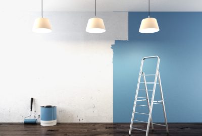 Room with a wall half-painted blue, a paint can, roller, and silver stepladder on a dark wood floor.