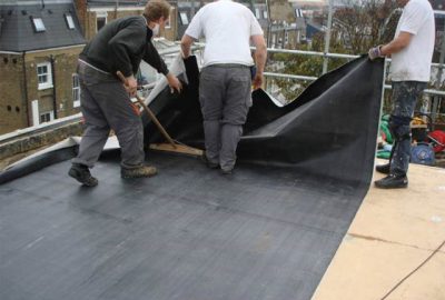 Three workers installing a black waterproof membrane on a flat rooftop.